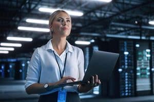 Female Data Center IT Technician Standing at the Server Rack Corridor with Laptop